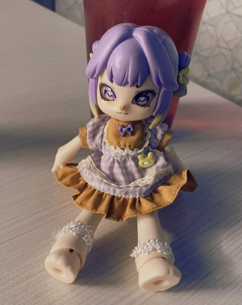 Lily, a rabbit-like doll with purple hair wearing a purple and orange dress sits against a cup of red juice.