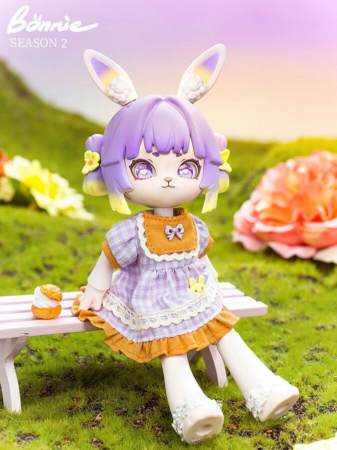Taro Lily is a small doll sitting on a set made to look like a bench outside. She has short, purple hair with yellow tips. Her bunny ears also purple and yellow. She wears a purple dress with orange accents. The top left of the image hax text: 'Bonnie Season 2.'