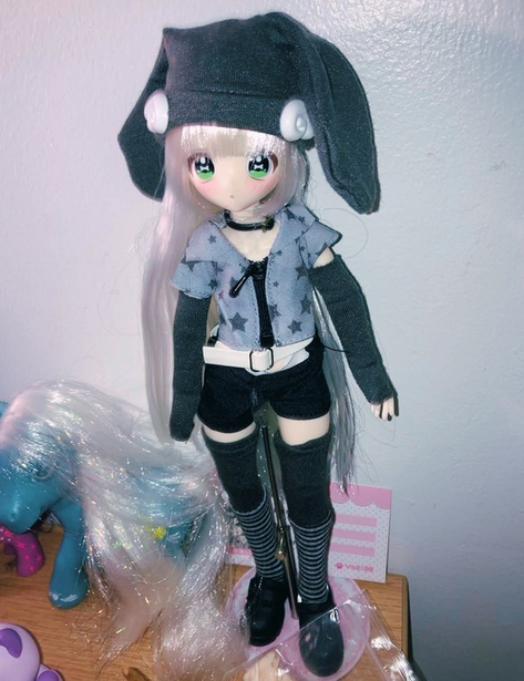 A blonde doll wearing a gray hat with large, floppy ears, a gray zip-up top with seperate gray arm warmers, black shorts with a white belt, and gray, striped socks. She is on a metal doll stand against a white wall. A blue gen 3 MLP stands besides her.